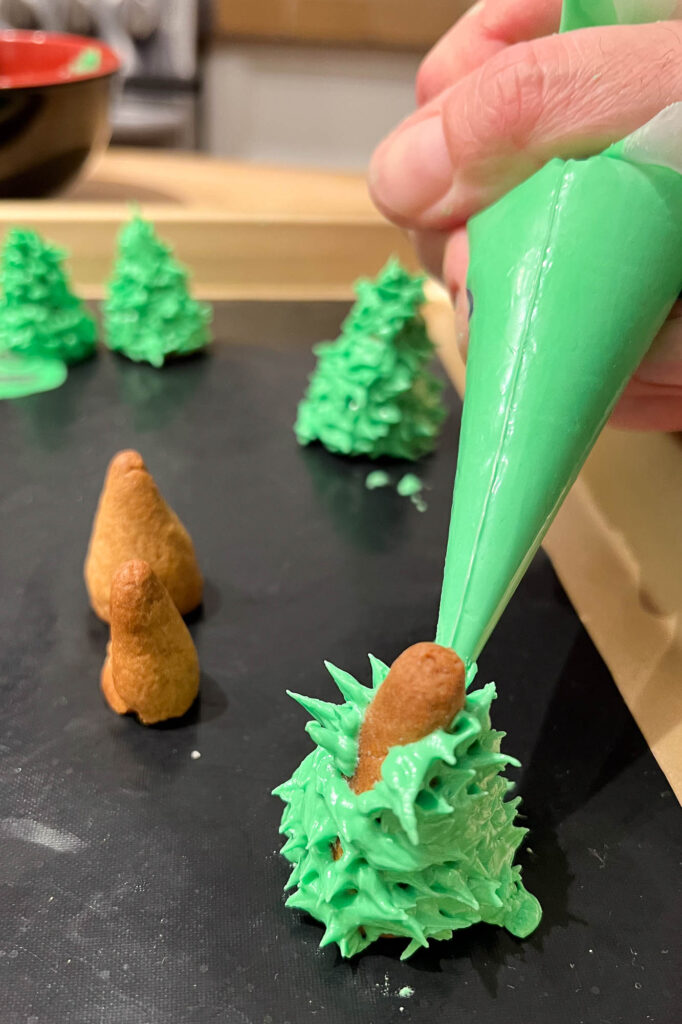 Green icing is being piped in the shape of fir trees.